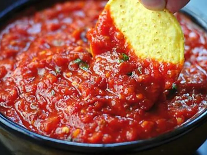 A bowl of chunky red roasted salsa with a tortilla chip being dipped into it