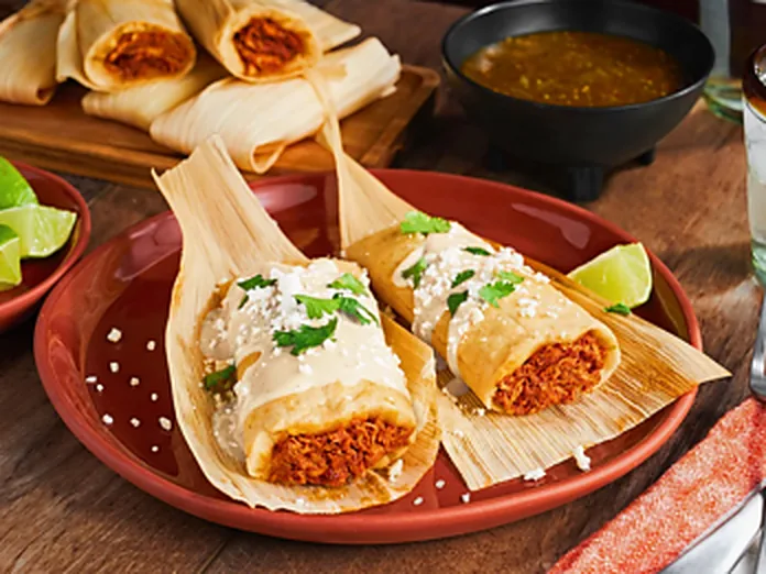 Tamales in corn husks served on a red plate with salsa and lime